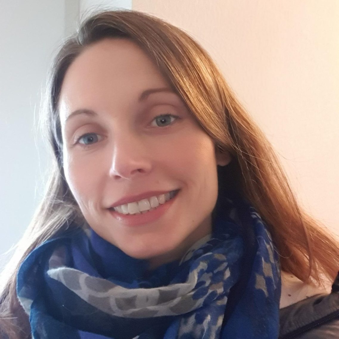 Lächelnde Frau mit langem, hellbraunen Haar und blau gemustertem Halstuch.